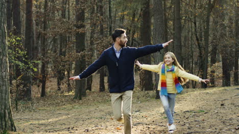 feliz padre caucásico y su hija corriendo en el bosque con las manos abiertas imitando un avión volando