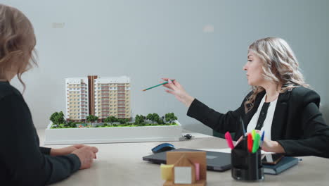 Business expert explaining details of architectural project to client while pointing at residential model, both engaged in serious discussion across office desk with focus on property