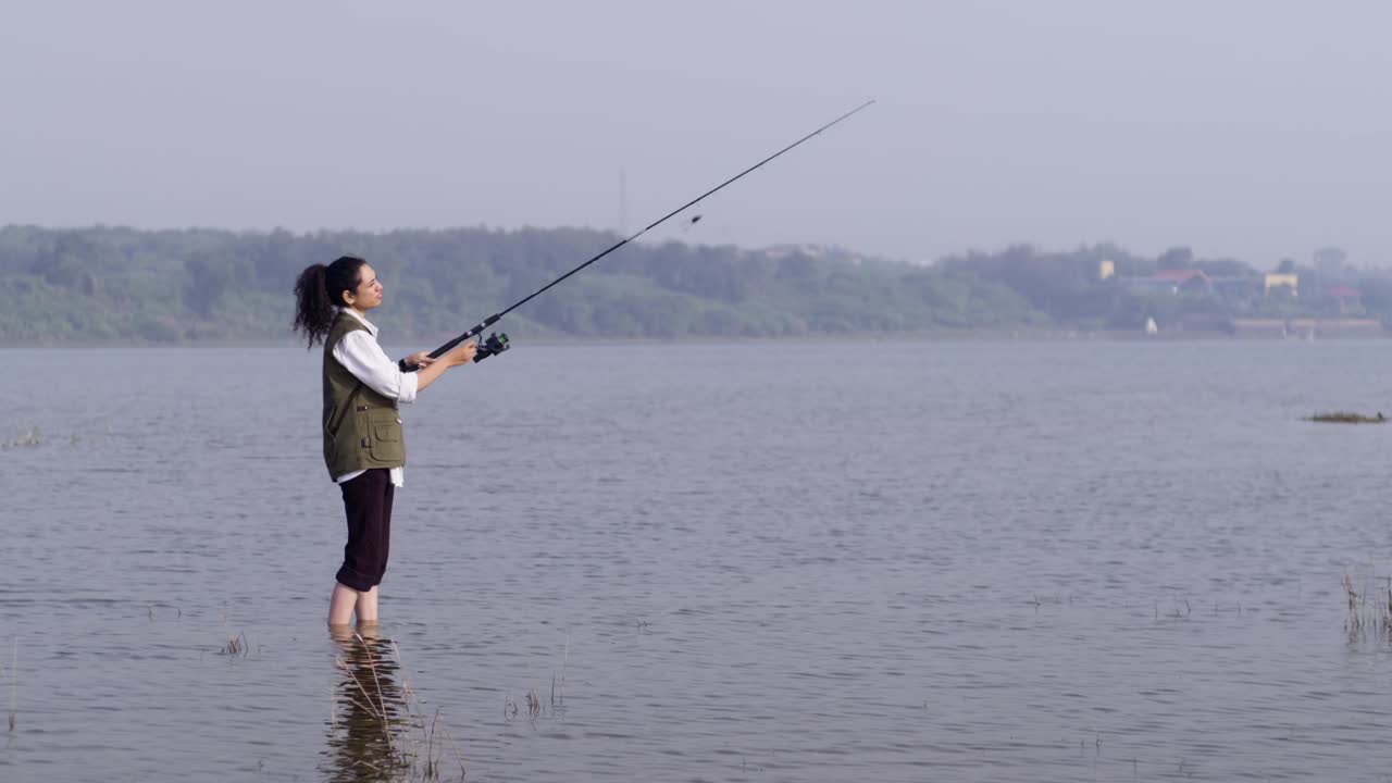 Seorang gadis Asia Selatan mengenakan rompi memancing berdiri di air danau yang dangkal, melemparkan pancingnya ke arah perairan terbuka