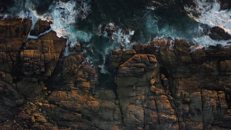 Foamy-Sea-Waves-Crashing-On-Rocky-Shore-At-Sunset---aerial-top-down