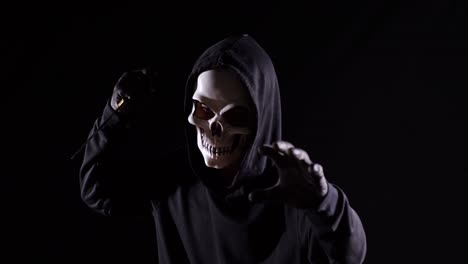 close up of scary man in the hooded sweatshirt wearing halloween mask holding a knife and making frightening gesture while walking on the black background