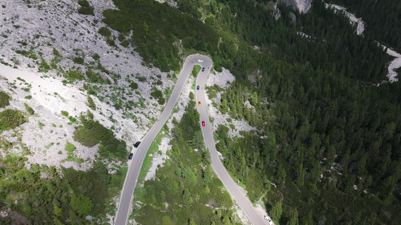 Drone shot of three exotic sports cars driving along a winding mountain road in the Alps, with forests and dramatic summer peaks in the background