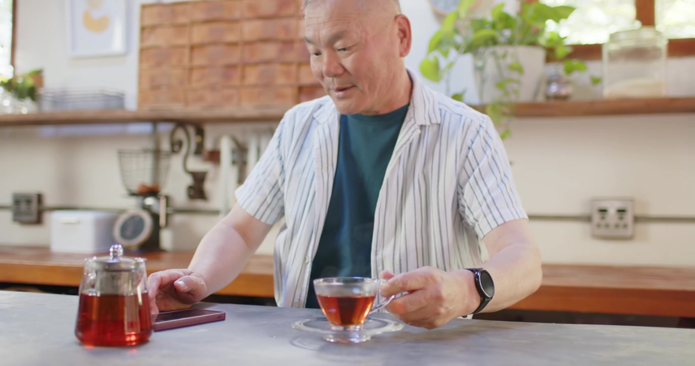 Anziano uomo asiatico al bancone della cucina che prende un tablet, toccando e scorrendo mentre sorseggia da una tazza di tè.