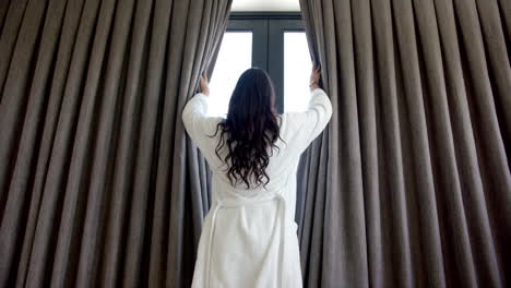 Rear-view-of-biracial-teenage-girl-drawing-bedroom-curtains-at-sunny-window,-slow-motion