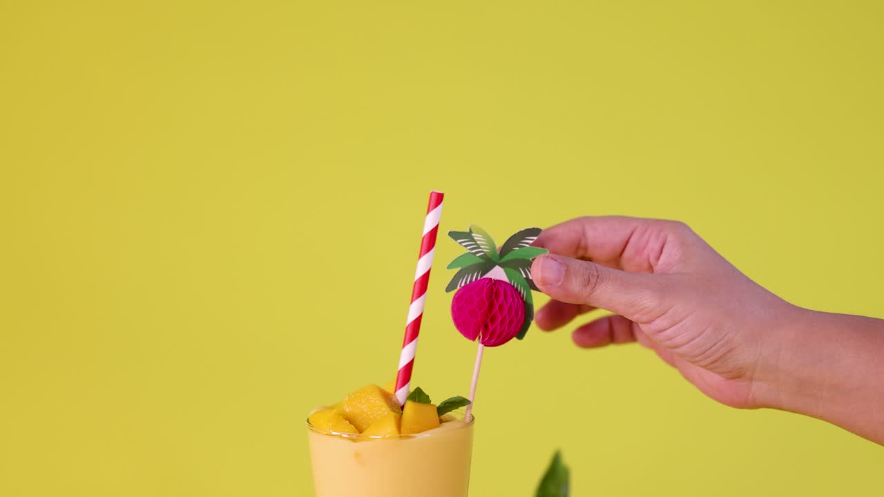 Hand adds paper fruit decoration to mango smoothie with fresh fruit, yellow background, bright lighting