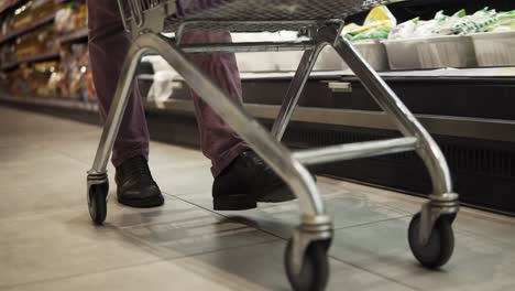 Shopping-in-the-supermarket,-view-of-the-male-legs-from-inside-the-trolley.-A-guy-pushing-shopping-cart-by-the-supermarket-aisle.-Cropped,-close-up-footage.-Slow-motion