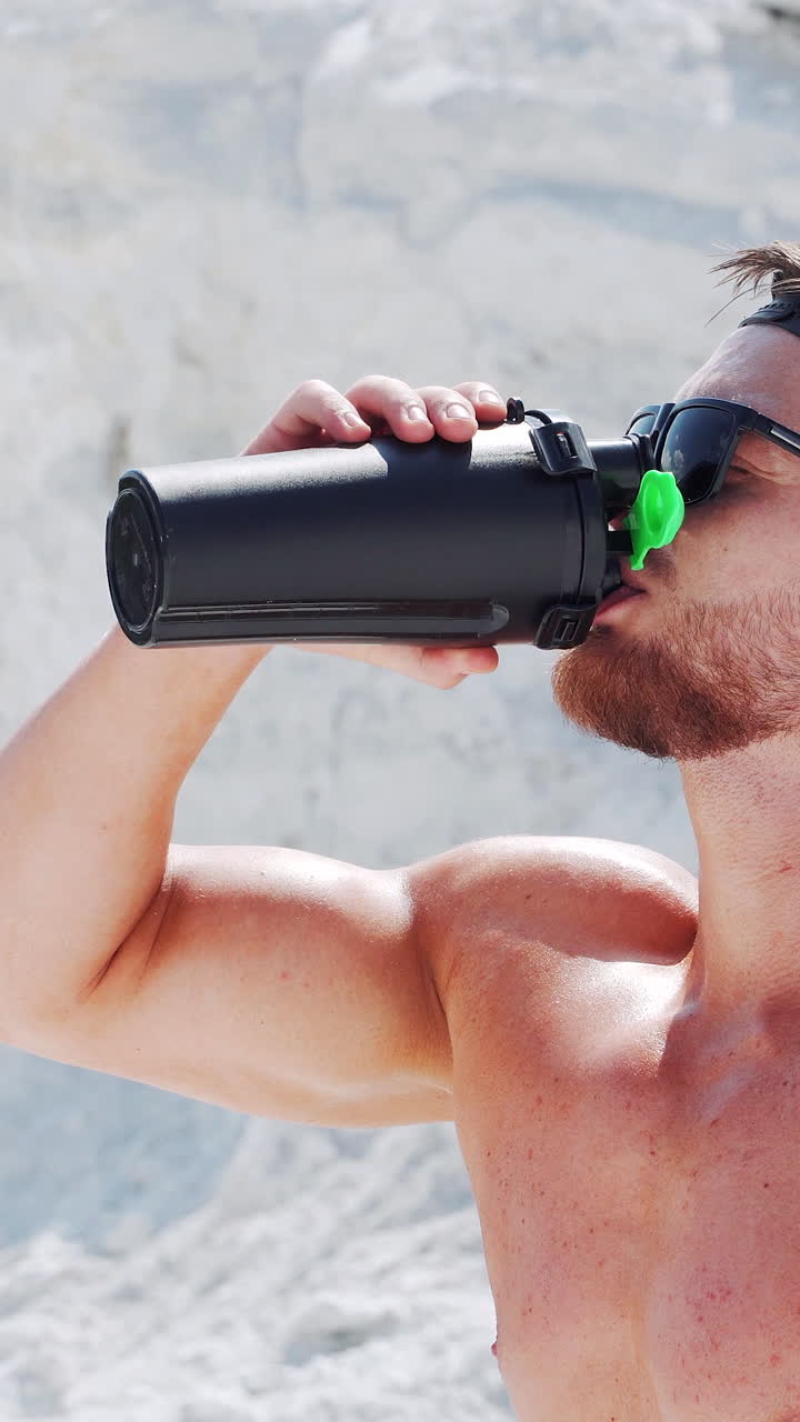 A strong naked man opens a black bottle and drinks water. Outdoor fitness. Vertical video