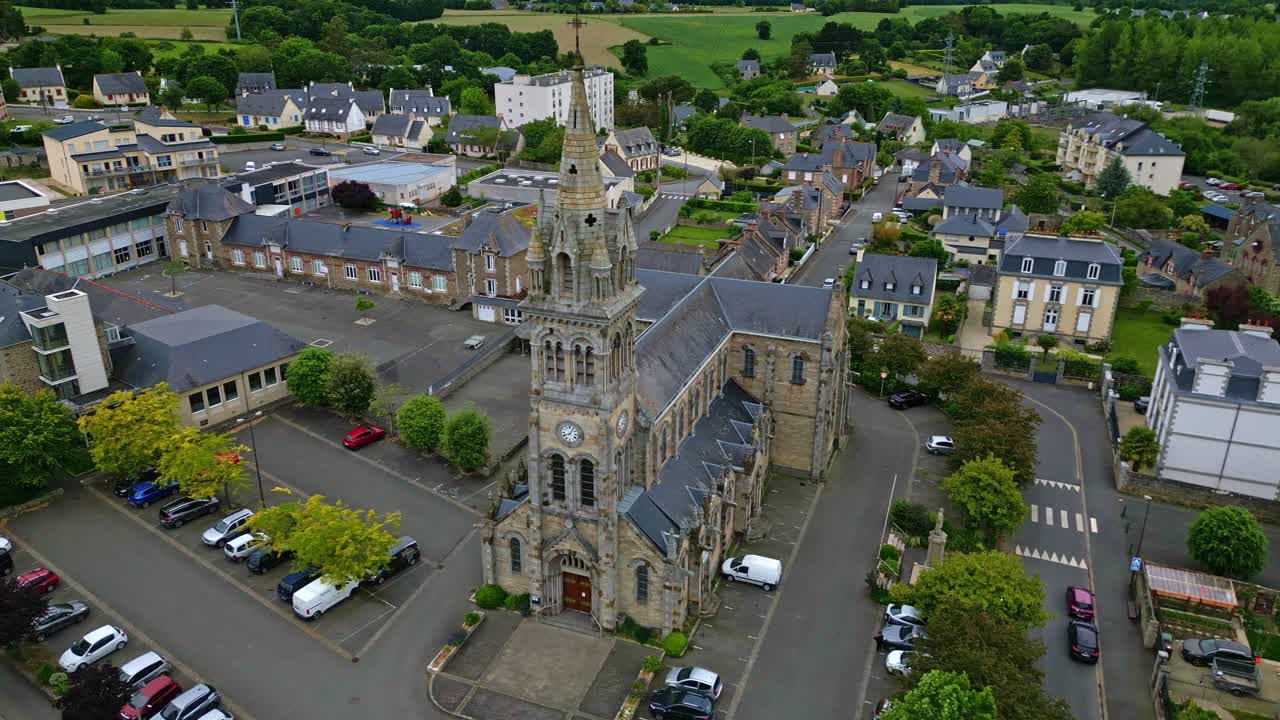 The drone flies laterally past Saint-Sauveur Church in Plancoët, revealing parked cars, a flying bird, nearby houses and green fields. Brittany in France