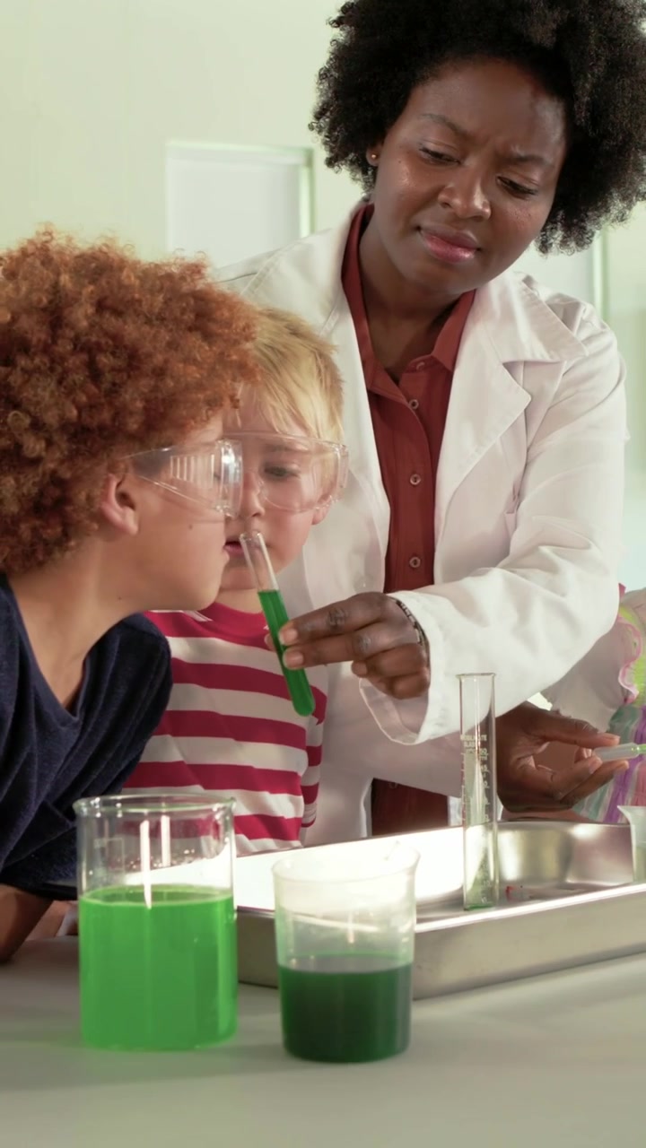 Vertical video: Lifting, teacher in coat guiding kids sniffing green test tube at bench, copy space