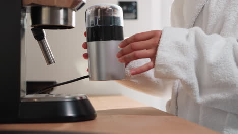 mujer moliendo granos de café en el dispositivo en la mesa de primer plano. dama en túnica de terry molienda granos en el molinillo en el hogar. procesamiento de producto de cafeína para hacer bebida