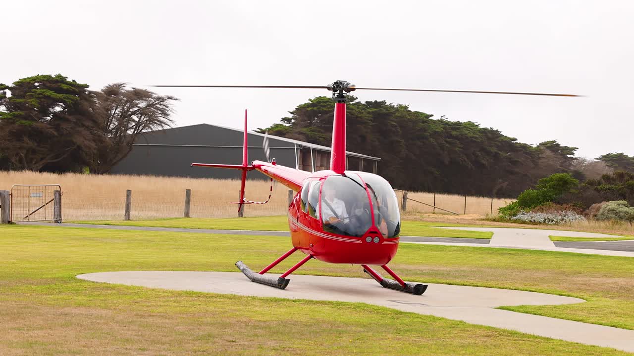 A red helicopter takes off from a grassy field in Port Campbell, Australia, under overcast skies, showcasing dynamic movement