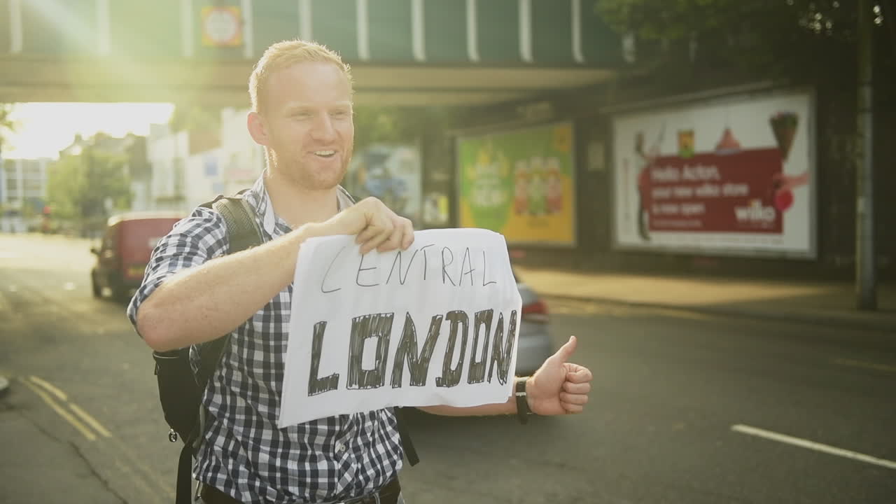 With a soft golden sunsetting glow surrounding him, a red-headed man holds up a sign hitchhiking to Central London