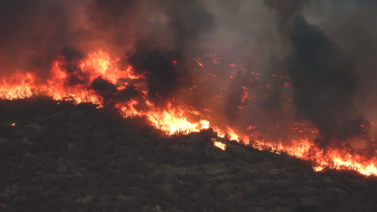 Incendio boschivo che brucia su un versante di montagna, con fumo nero che si innalza.