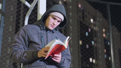 man in hoodie and beanie leaning against metallic goalpost reading red book outdoors at night with mesh fence and illuminated urban city background, thoughtfully flipping pages