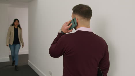 male teacher talking on mobile phone passing female colleague as they walk along corridor in school building