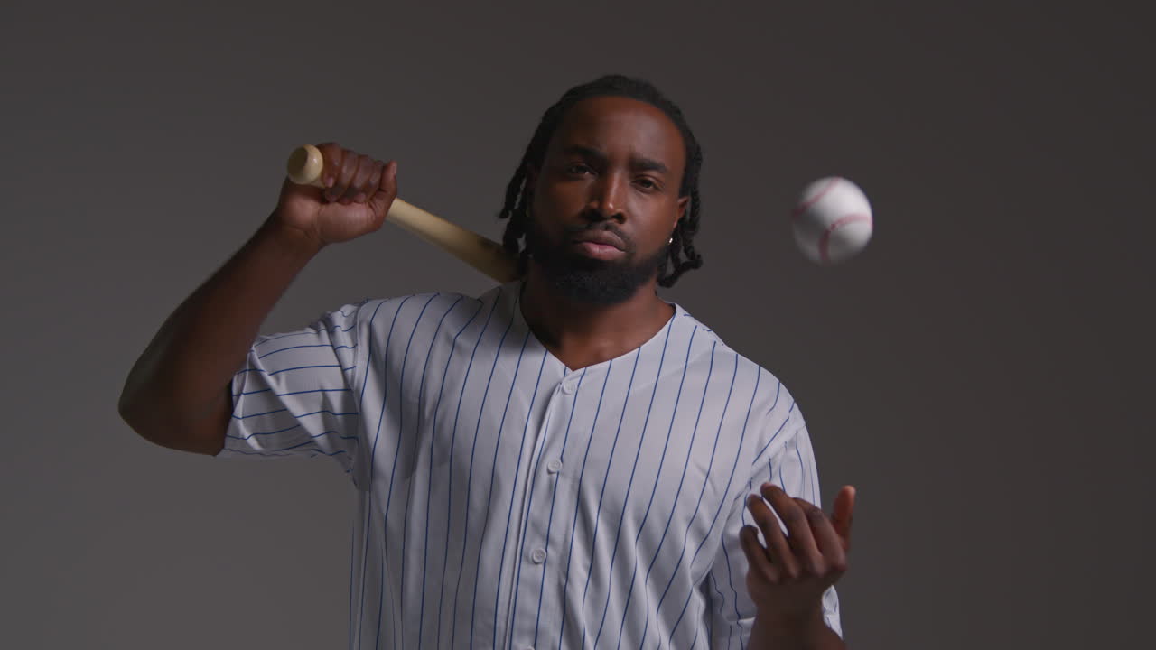 Studio Portrait Of Serious Male Baseball Player Wearing Team Shirt Holding Bat And Throwing Ball In The Air Shot Against Grey Background