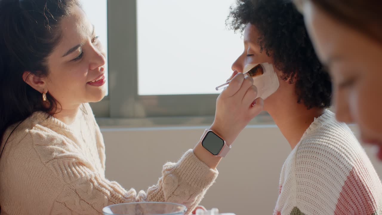 Diverse female friends dipping brush into mask bowl, applying mask and painting nails for home spa