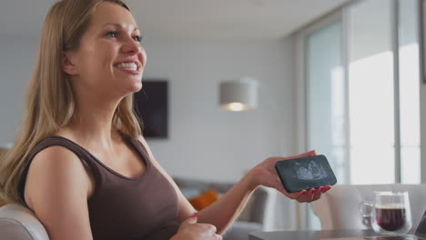 mujer embarazada sonriente en casa sosteniendo un teléfono móvil con ecografía del bebé