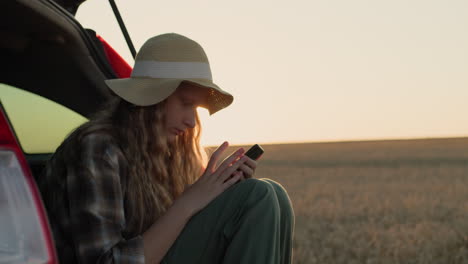 a teenage girl sits in the trunk of a car, uses a smartphone. against the backdrop of a rural landscape where the sun sets