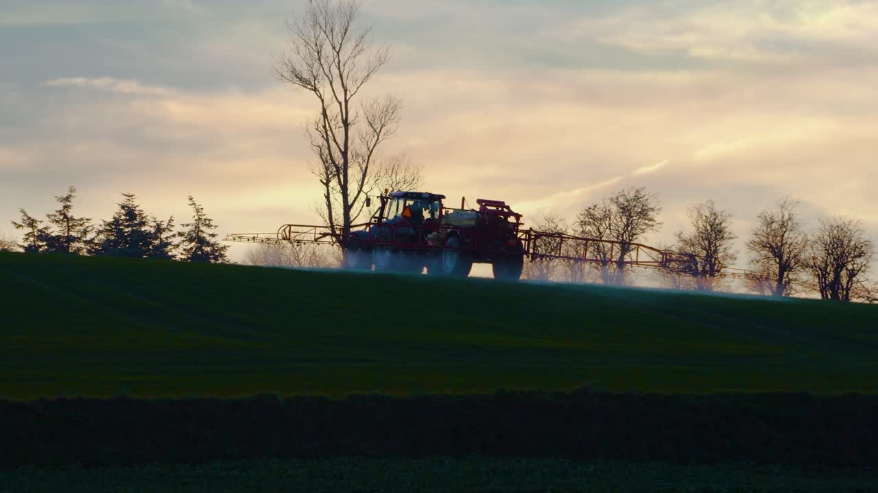 A farmer with a tractor spraying chemicals to a field during sunset