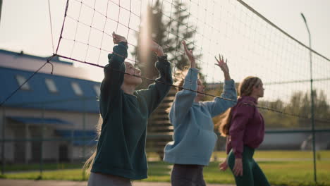 females in hoodies engage in volleyball training outdoors on court, background features, greenery, and building, women jump with hands raised during practice session