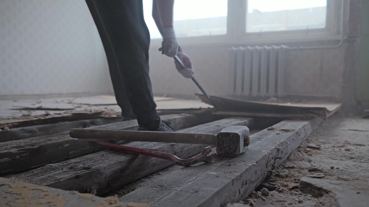 Low angle reveals heavy tools as worker lifts damaged flooring through dusty air