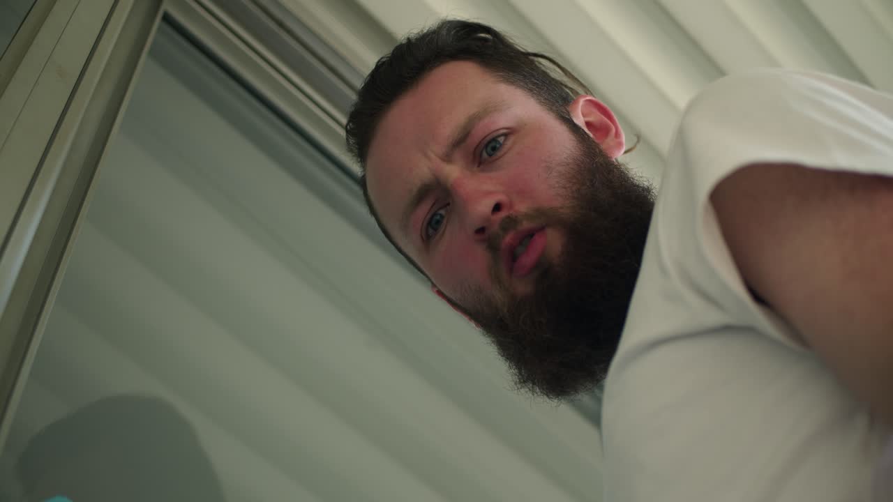 A low-angle shot captures a man with a beard and a white shirt, his face tense and curious as he looks downward. The perspective evokes a sense of unease or confrontation.