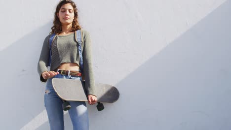 Happy-mixed-race-woman-holding-skateboard,-leaning-against-wall-smiling-in-the-sun