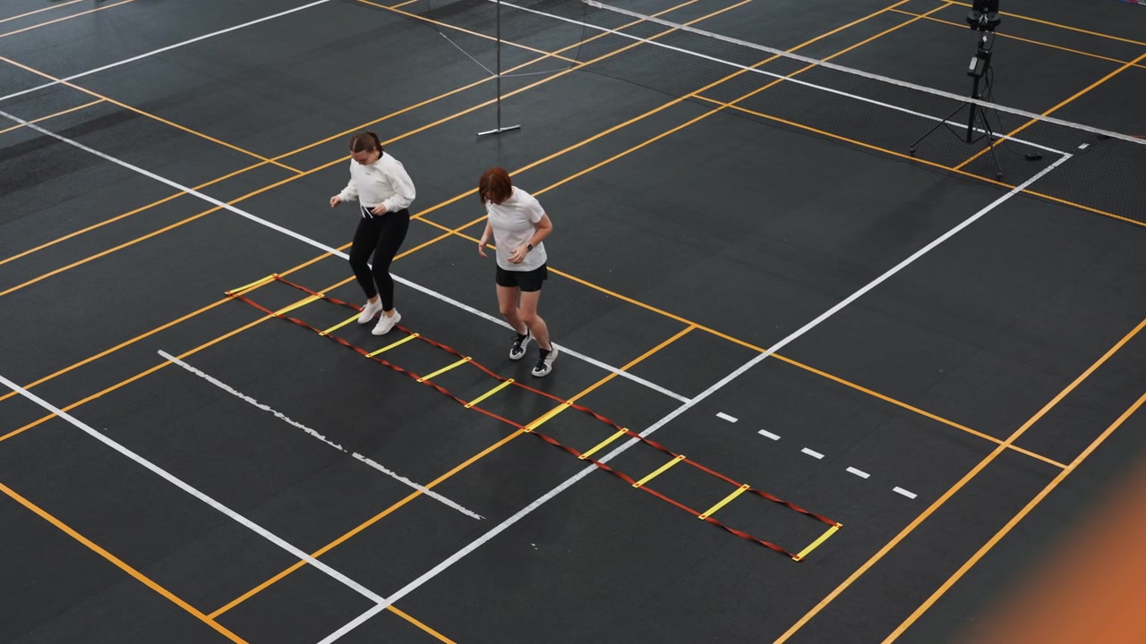 White therapist evaluates badminton footwork on court ladder during clinical assessment, measured steps and gait observation in indoor training hall, focus on mobility strength and injury prevention