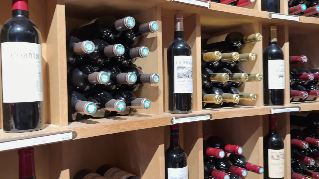 Static medium shot of numerous red wine bottles organized on light wooden cellar shelves