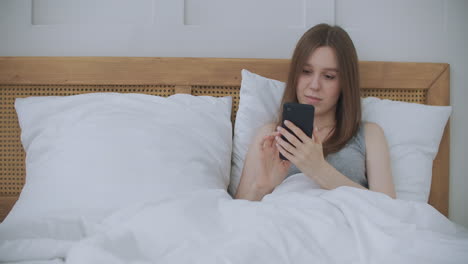 vista de cerca de una mujer escribiendo un mensaje en un teléfono móvil con 2 manos mientras está cómodamente acostada en una cama en una habitación de hotel. mujer usando un teléfono inteligente y acostada en la cama por la mañana después de despertarse