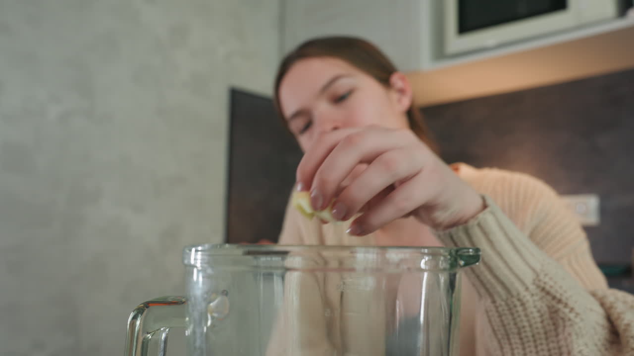 Mujer embarazada preparando bebida en licuadora tranquilamente, iluminación interior suave, suéter acogedor, jarra de licuadora de vidrio en la encimera, mano suave añadiendo fruta o suplemento, rutina prenatal tranquila enfatizando lo maternal