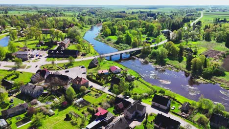 a tranquil river flows under a connecting bridge, bordered by charming houses and lush green surroundings in biržai, lithuania - orbit drone shot