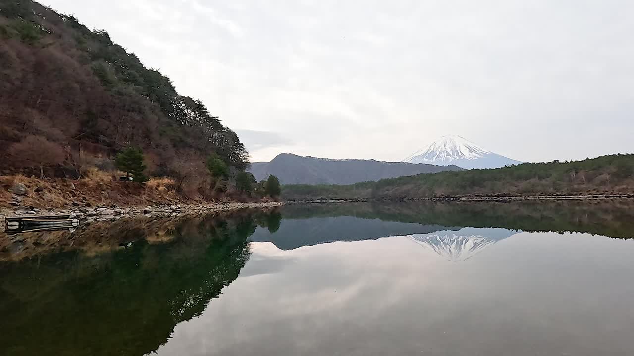 Mount Fuji reflects on the tranquil waters of Lake Saiko under overcast skies, capturing a peaceful and scenic Japanese landscape