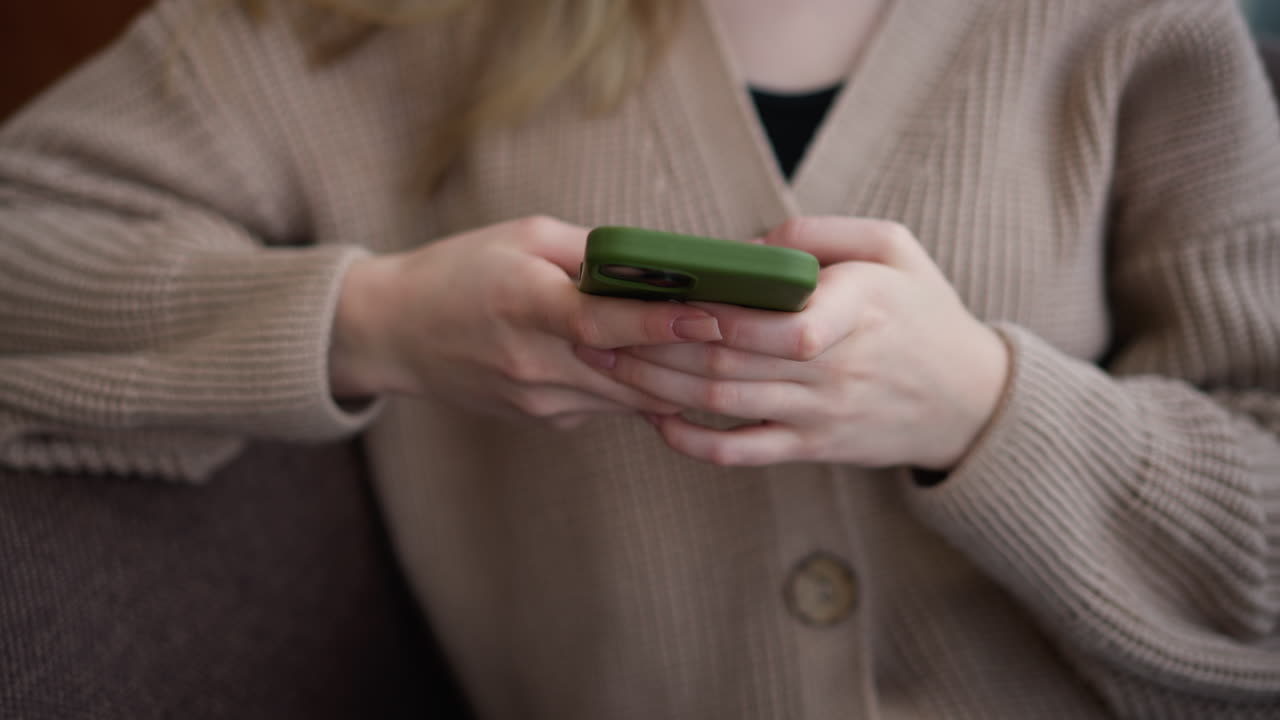 나무 테이블에 앉아있는 젊은 여성이 녹색 케이스로 아이폰을 작동하는 클로즈업, 그녀의 손은 스크린에 초점을 맞추고, 편안한 환경에서 세부 사항에 주의를 기울이고 있습니다.