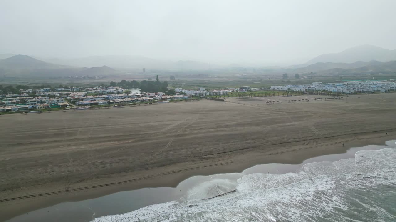 Drone footage of a beach in the morning, with waves crashing on the shore and beach houses in the distance, all under a misty, cloudy atmosphere.