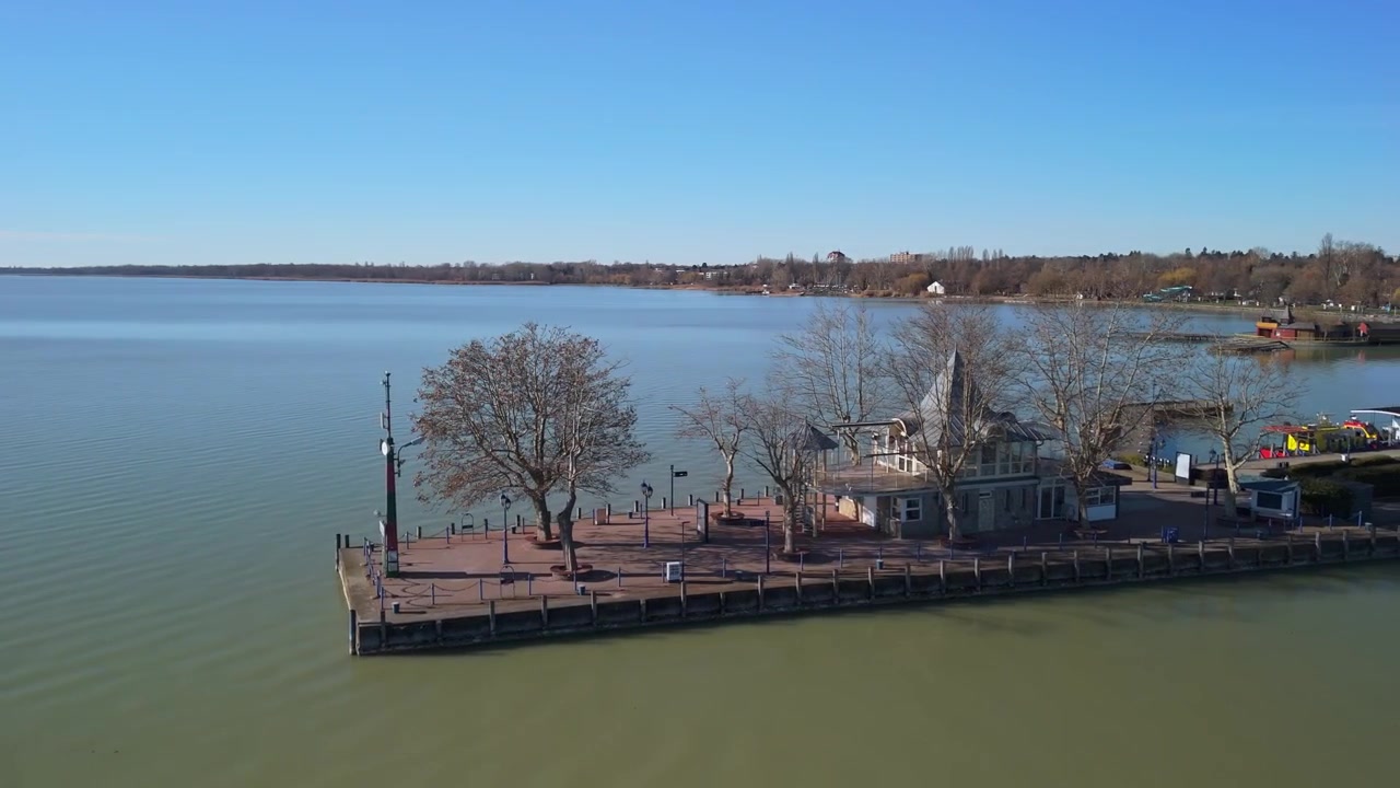 I Keszthely viser et flyfoto marinaen langs den rolige strandlinjen ved Balatonsjøen, med bladløse trær og historiske bygninger som grenser til den rolige vannkanten under en klar himmel.