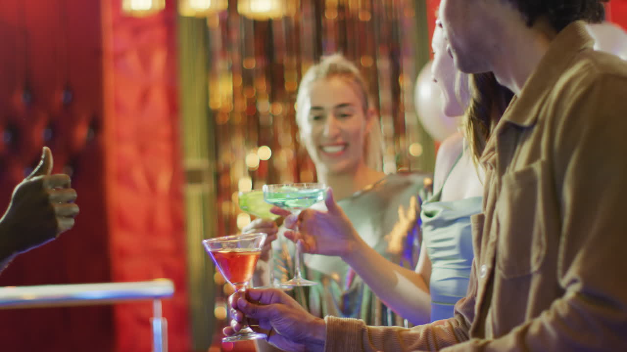 Video of diverse group of happy male and female friends drinking cocktails standing at a bar