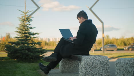 freelancer seated outdoors working on laptop with leg crossed, surrounded by greenery and urban environment, background features parked cars on opposite lane, moving car, and modern structures