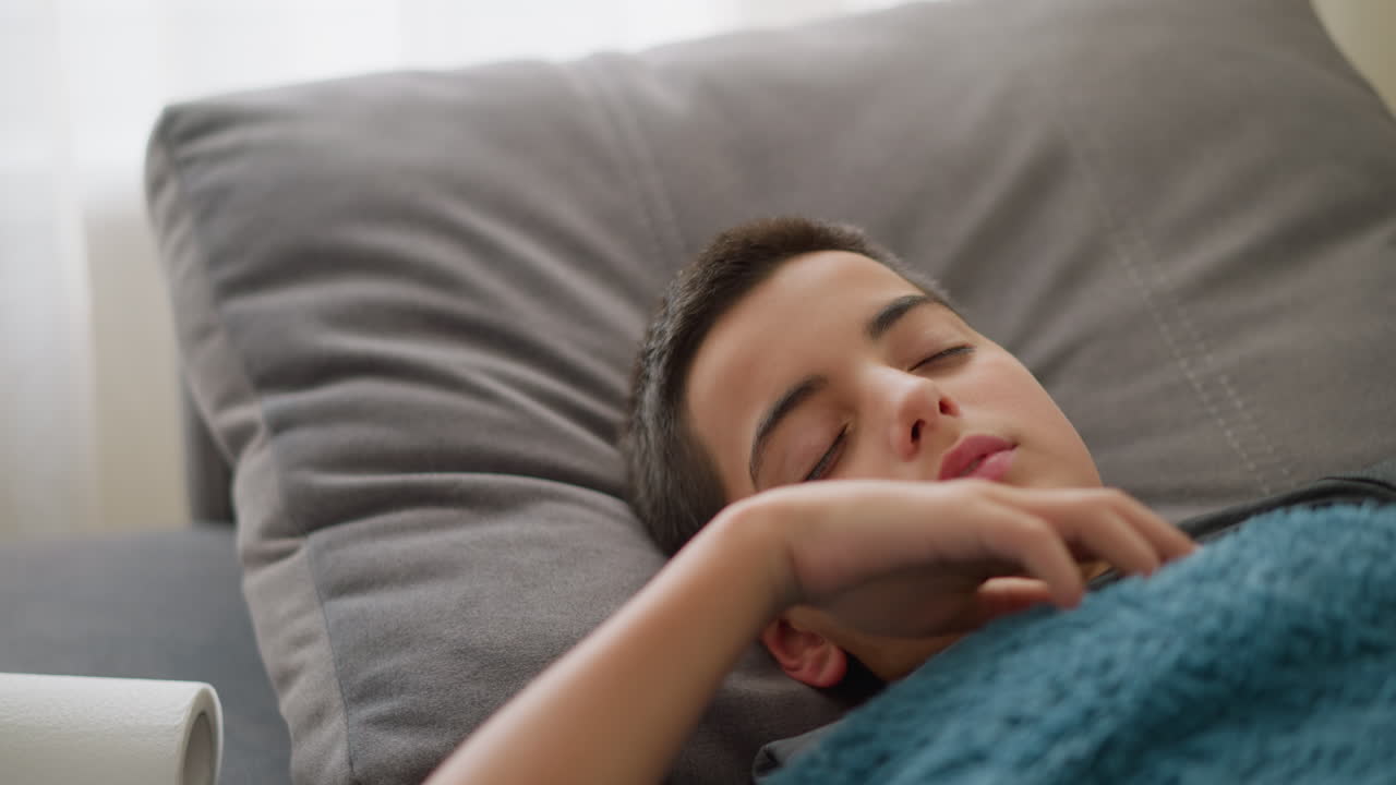 Close-up of sick child resting on bed covered with blue blanket, turning with discomfort while coughing, with tissue nearby and light streaming through window