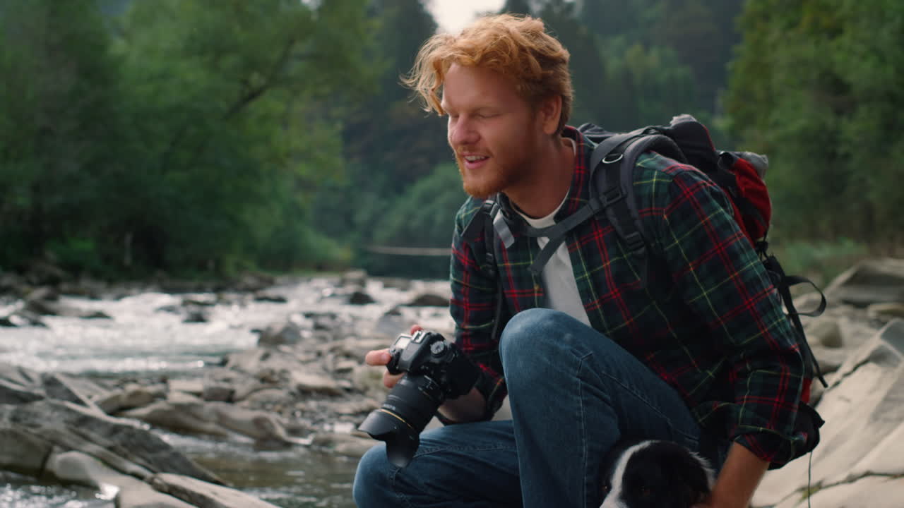 fotógrafo sentado en la orilla del río con el perro. tipo tomando fotos en la cámara