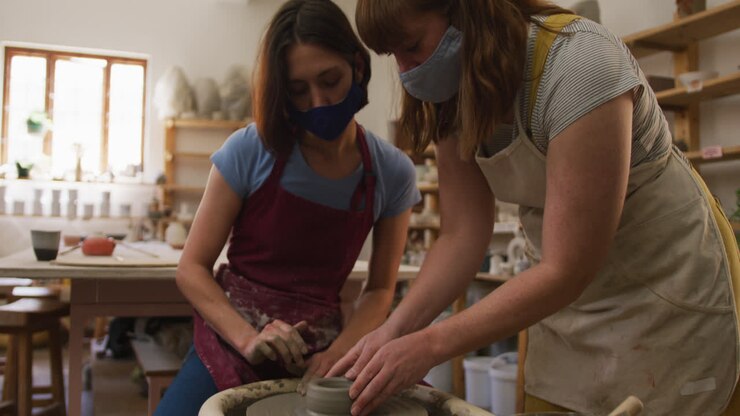 Two female caucasian potters wearing face masks and aprons creating pottery on potters wheel at pott