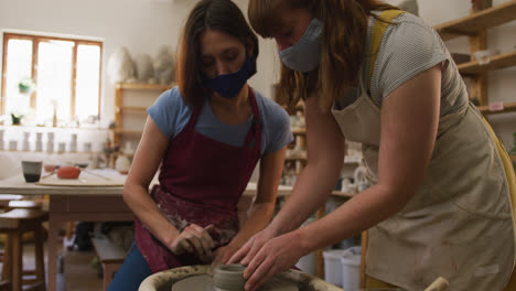 two female caucasian potters wearing face masks and aprons creating pottery on potters wheel at pott