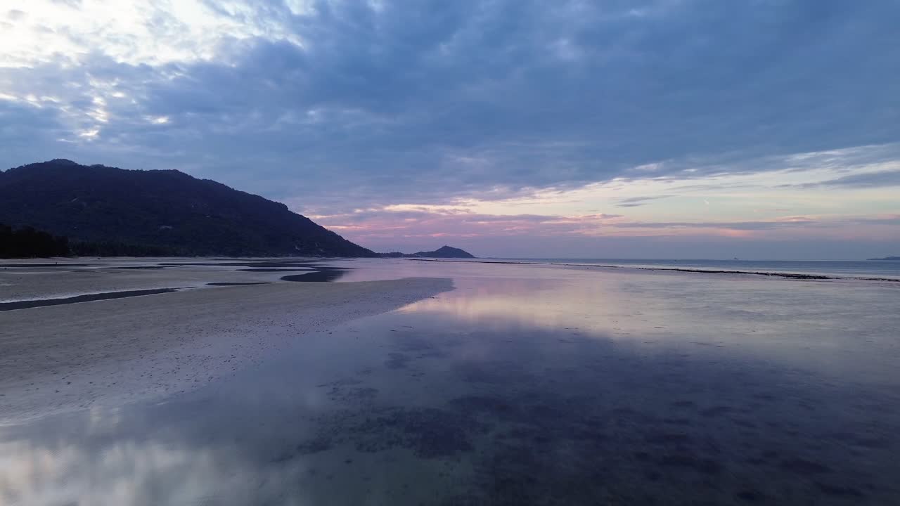 Beautiful aerial footage of sunrise over Koh Phangan in Thailand with calm ocean and pink sky colors reflecting on the beach during low tide