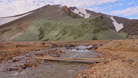 Amplia-Vista-Fija-Del-Río-Que-Desciende-Desde-Brennisteinsalda-En-Landmannalaugar,-Islandia,-Con-Un-Pequeño-Puente-De-Madera-Para-Excursionistas-Frente-A-La-Montaña-Bláhnúkur.