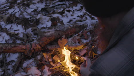 close-shot-looking-over-the-shoulder-of-a-bearded-man-who-is-adding-small-sticks-and-kindling-to-a-newly-lit-campfire