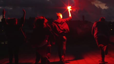 un grupo de adolescentes felices con una luz de señal sostenida y agitando caminando hacia adelante hacia afuera cámara por la noche.