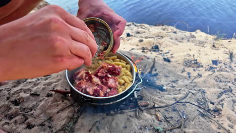 Slow motion close-up shot of person scraping fish meat out of tine can with pocket knife into pasta dish camping cookware pot food meal cooking on fire
