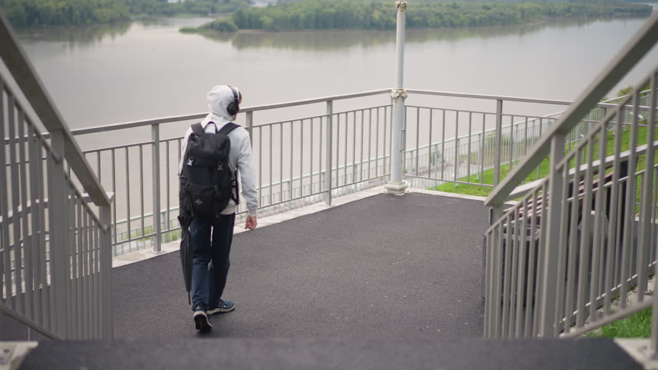 강가의 강을 내려다보는 검은 옷을 입은 남자가 후드와 헤드폰을 조절하고, 흐린 하늘 아래 금속 난간에서 잠시 멈춰 서서 우산을 꽂고, 잔잔한 물과 멀리 보이는 수평선, 침울한 분위기, 역할