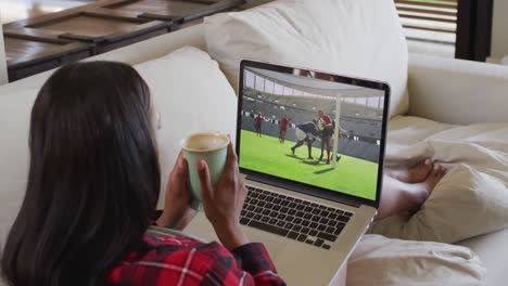 Composite-of-woman-sitting-at-home-holding-coffee-watching-rugby-match-on-laptop